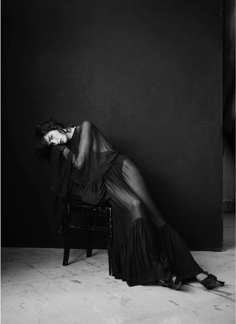 A woman in a sheer black dress reclines on a chair in a black and white fashion shot with low-key lighting against a dark backdrop.