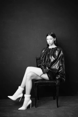 Black and white portrait of a woman in a dark shiny shirt and white heeled boots, sitting on a chair against a dark backdrop.