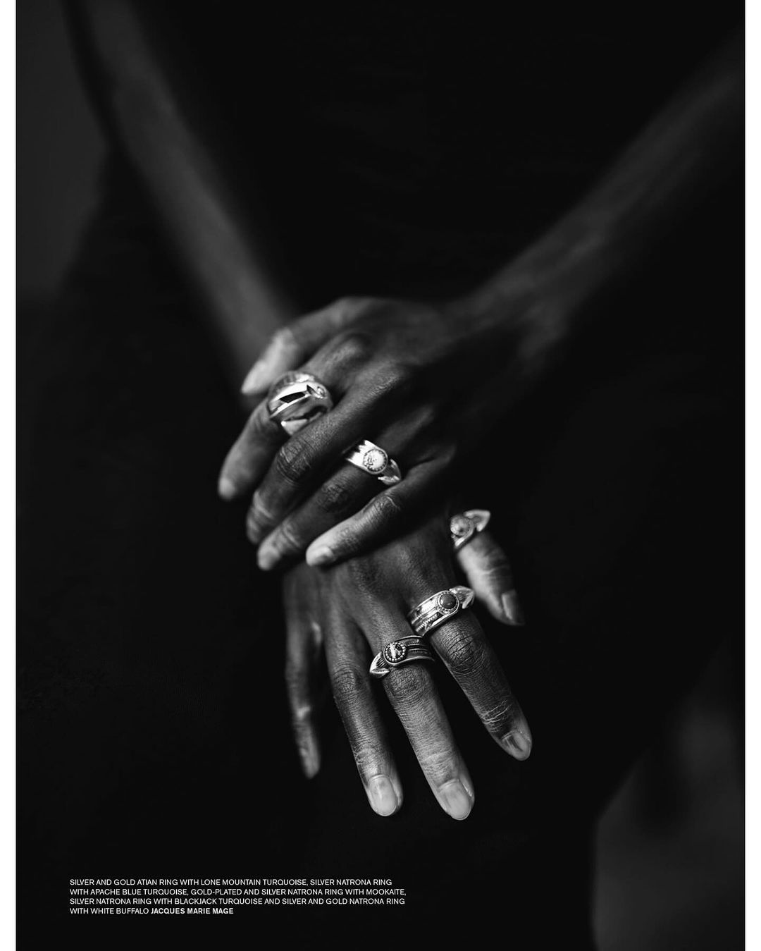 Close-up of dark-skinned hands adorned with several statement rings, set against a dark background highlighting jewelry.