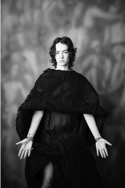 Monochrome portrait of a woman in a black avant-garde dress with arms outstretched against a painted backdrop in a studio setting.