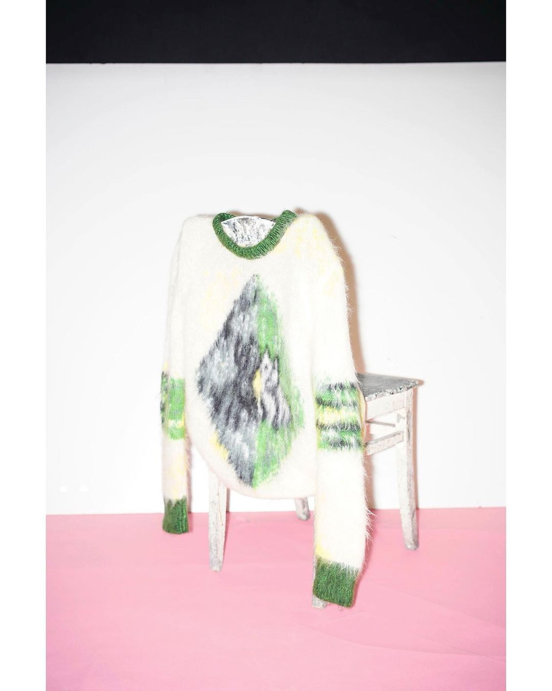 Fluffy white sweater with green trim and an abstract mountain design, displayed on a small chair against a pink and white background.