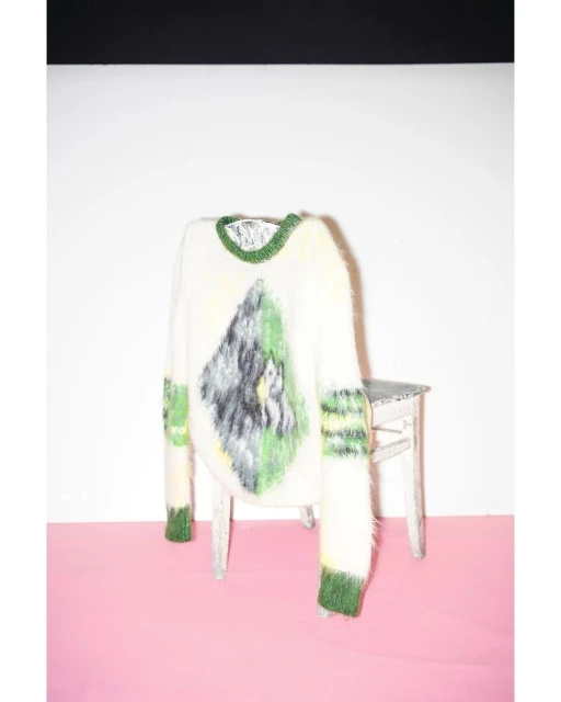 Fluffy white sweater with green trim and an abstract mountain design, displayed on a small chair against a pink and white background.
