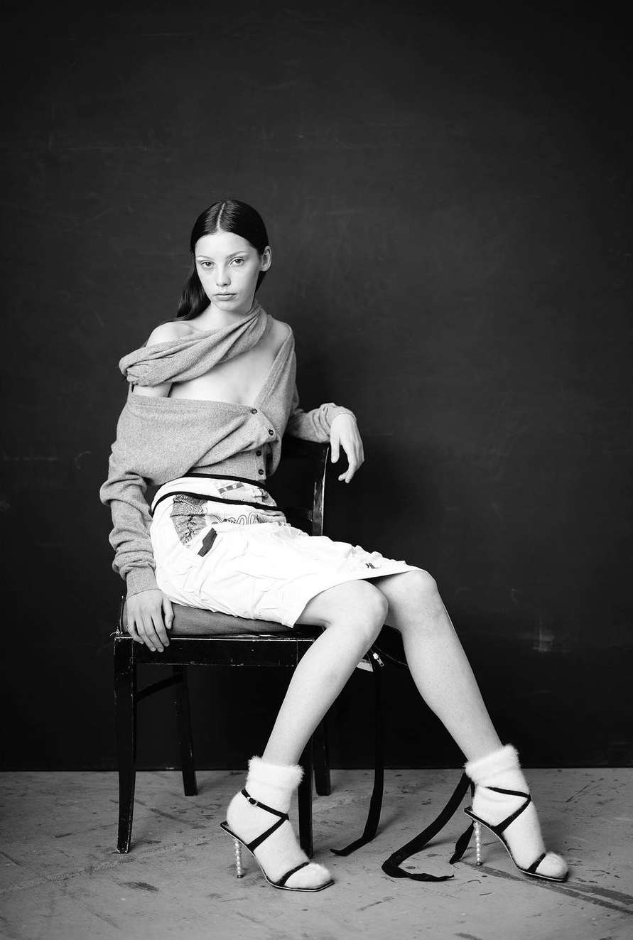 Black and white portrait of a young woman in casual fashion, seated on a chair against a dark backdrop in a studio setting.