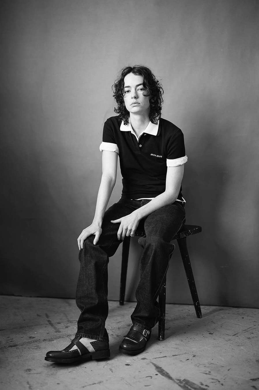 Monochrome portrait of a young adult sitting on a wooden stool, wearing a black polo shirt, jeans, and shoes against a gray backdrop.
