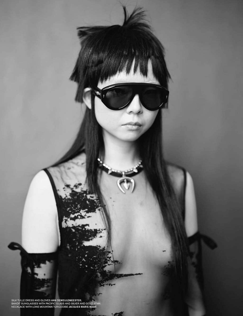 Monochrome portrait of an Asian woman with an elaborate hairstyle, wearing sunglasses and a necklace, against a gray backdrop.