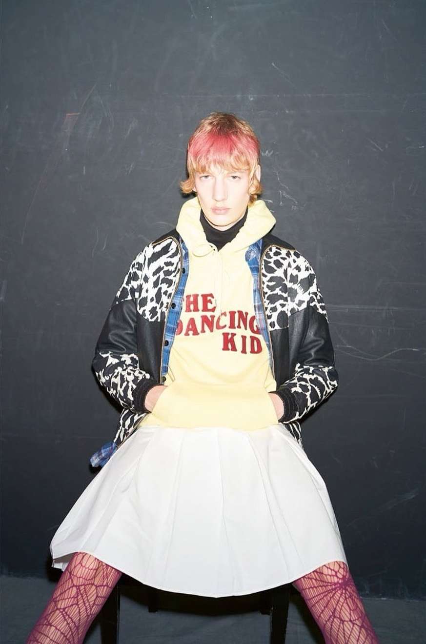 A fair-skinned person with pink hair poses in a yellow hoodie, cow-print jacket, white skirt, and patterned tights against a dark wall.