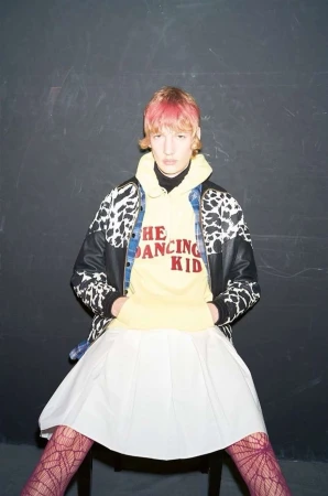 A fair-skinned person with pink hair poses in a yellow hoodie, cow-print jacket, white skirt, and patterned tights against a dark wall.