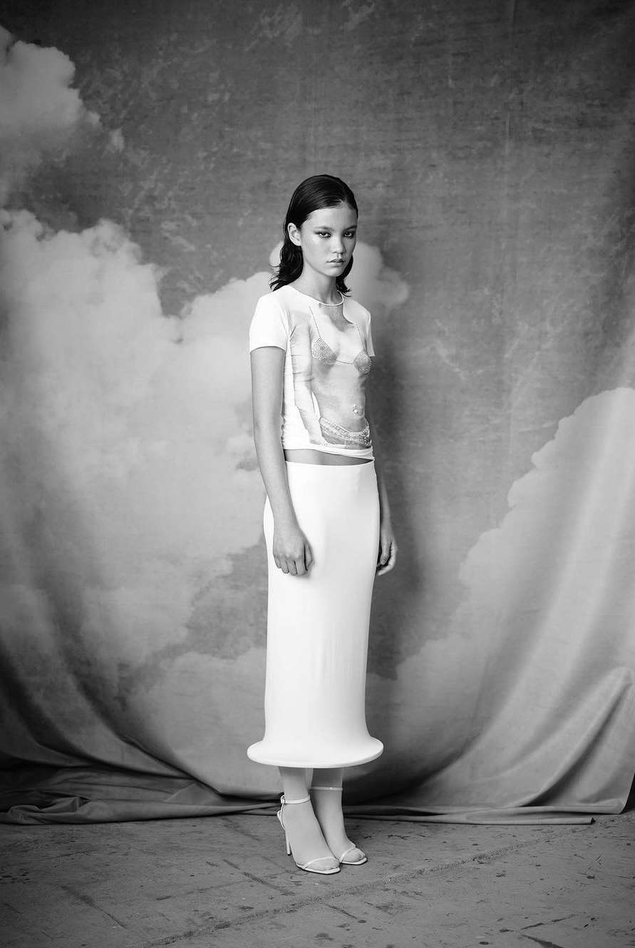 Black and white fashion shot of a woman in a white t-shirt with image graphic and long skirt against a cloud-patterned backdrop.
