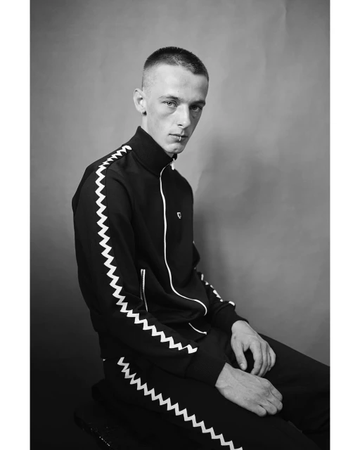 Monochrome portrait of a slender adult male in a black tracksuit with white chevron detail, seated against a plain grey background.