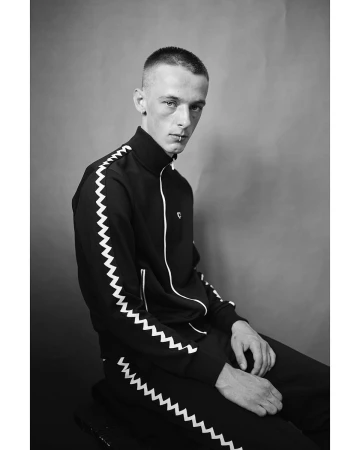 Monochrome portrait of a slender adult male in a black tracksuit with white chevron detail, seated against a plain grey background.
