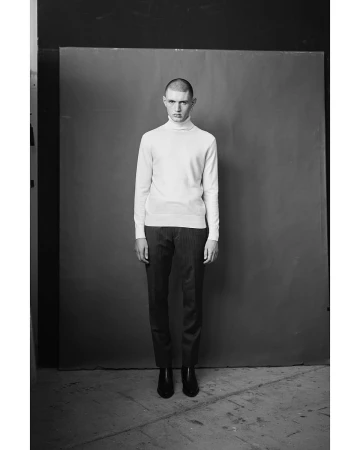 Monochrome portrait of a young man wearing a white turtleneck sweater and dark trousers standing in a studio.