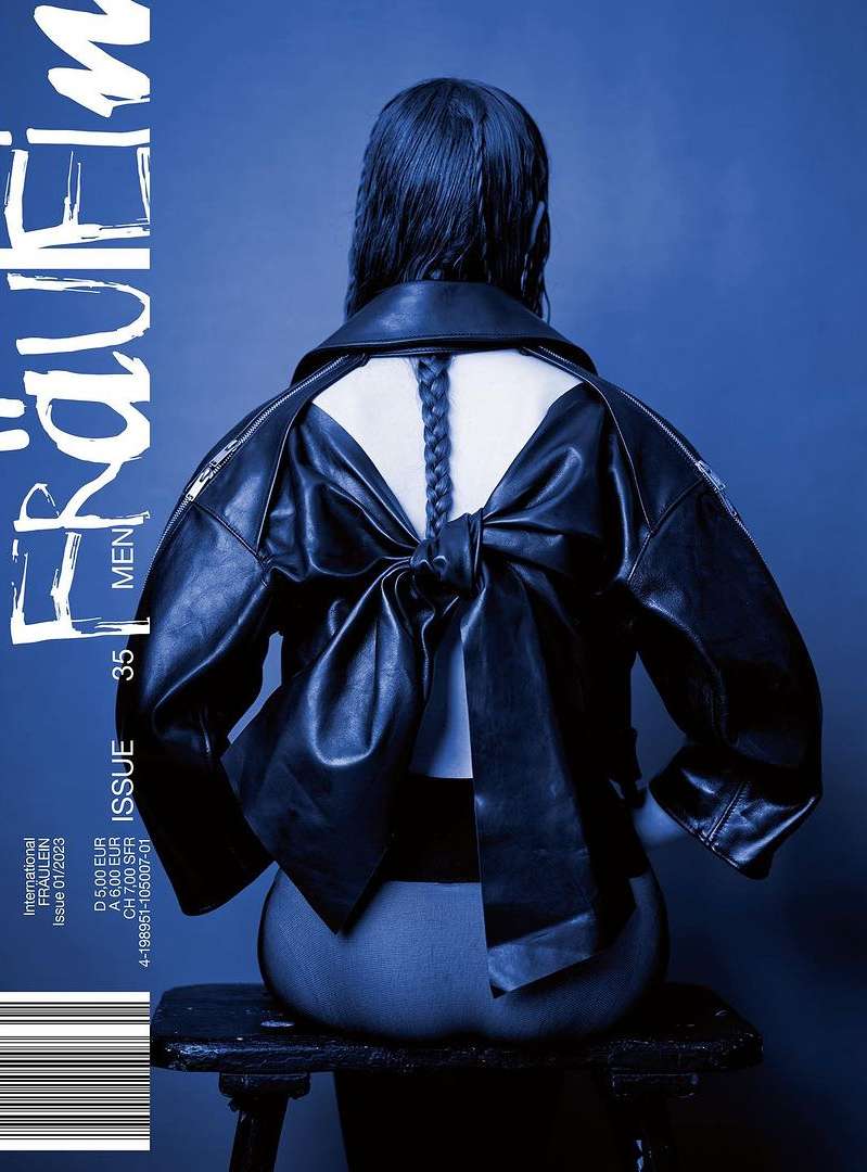 Back view of a woman with a braided hairstyle sitting on a wooden stool, wearing a black leather jacket with a bow detail.