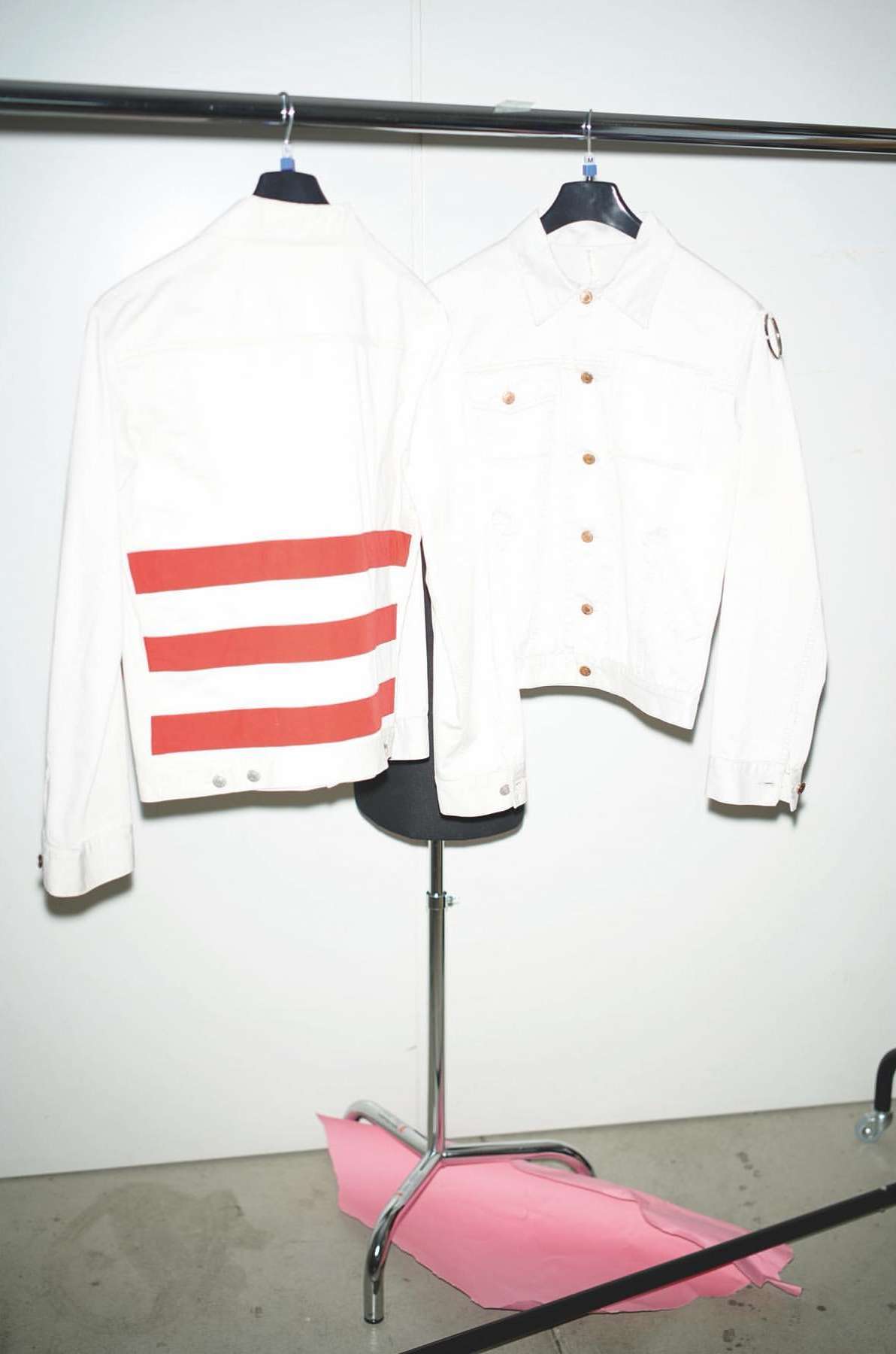 Two white jackets hang from a metal rack, one with red stripes, against a white wall in an indoor environment with natural light.