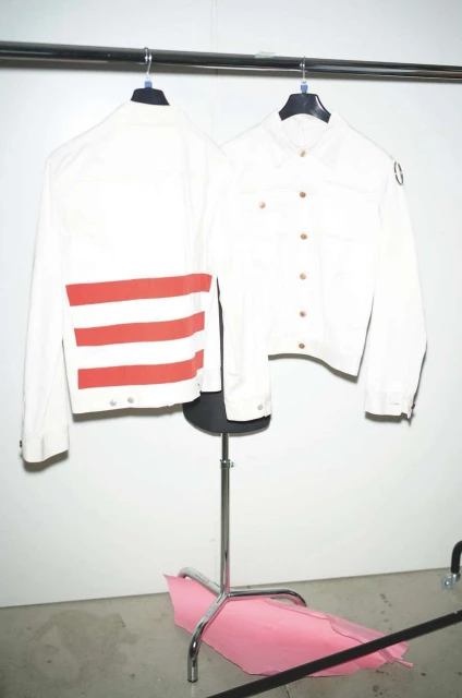 Two white jackets hang from a metal rack, one with red stripes, against a white wall in an indoor environment with natural light.