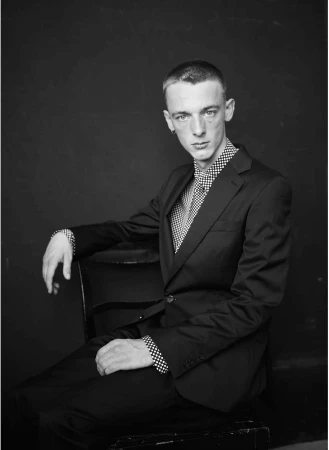Monochrome portrait of a young man in a dark suit jacket and patterned shirt seated against a dark textured backdrop.