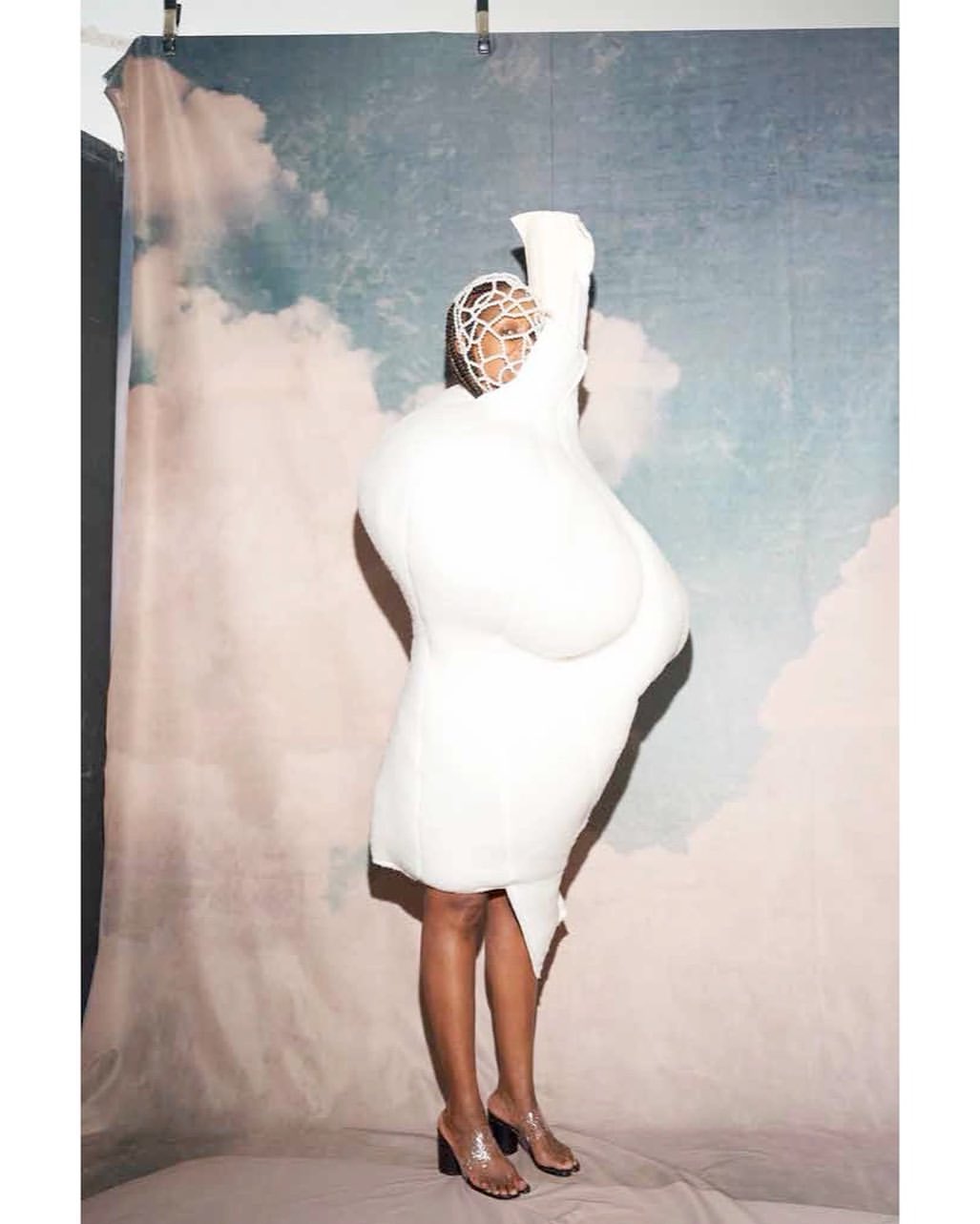 Fashionable adult female posing in studio wearing a unique voluminous white dress and transparent high heels in a fashion pose.