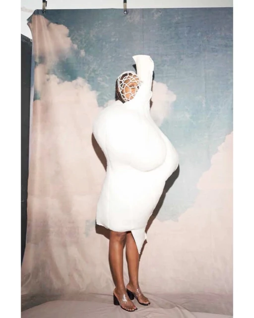 Fashionable adult female posing in studio wearing a unique voluminous white dress and transparent high heels in a fashion pose.