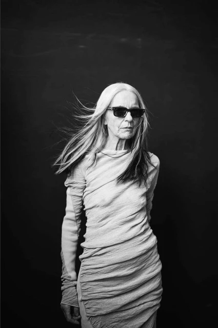 Black and white portrait of an older woman with long gray hair and sunglasses, wearing a draped dress against a dark studio background.