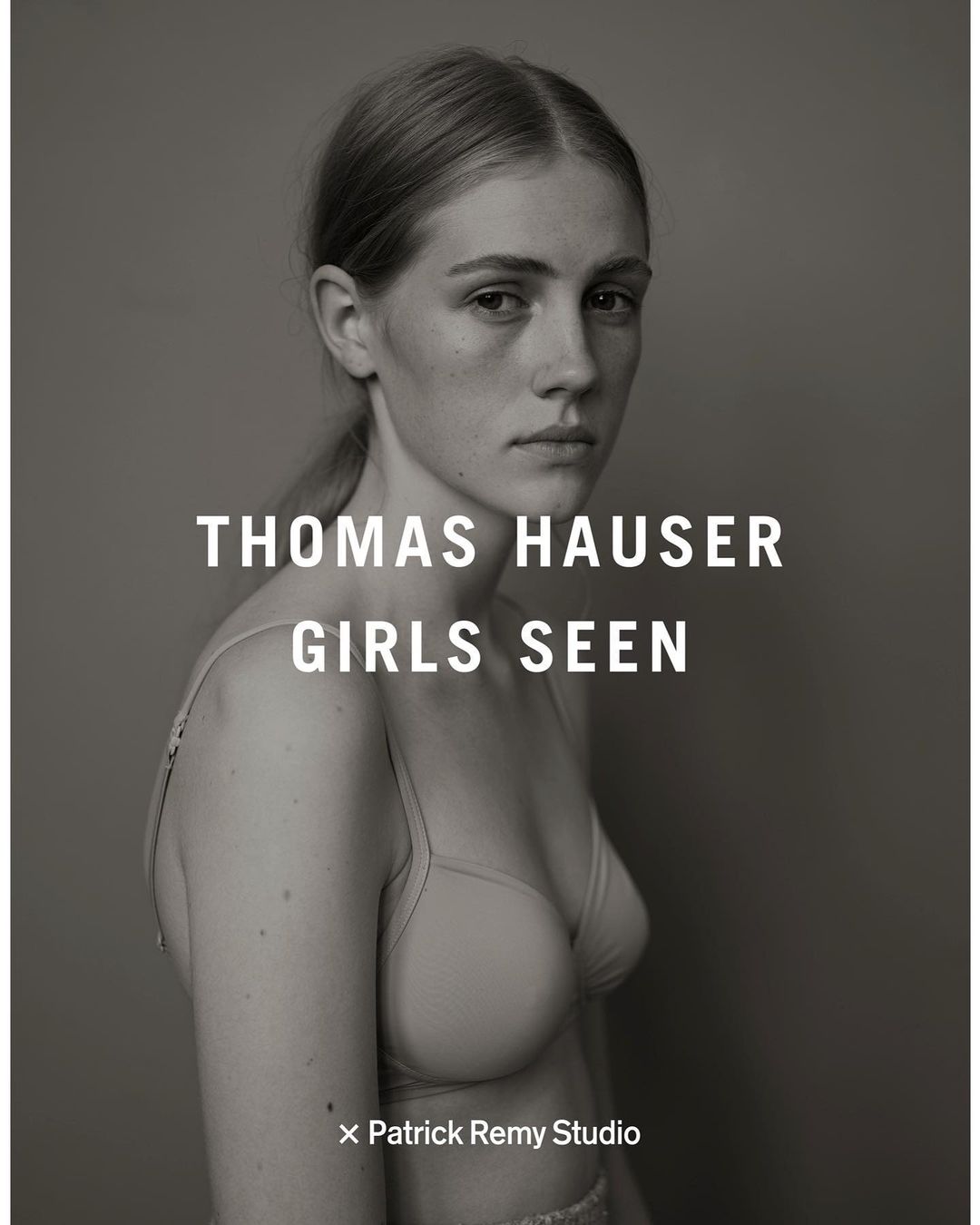 Black and white portrait of a young woman with blond hair looking at the camera, wearing a bra, posing in front of a grey background.