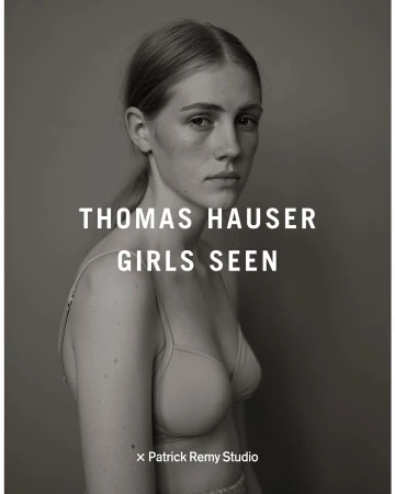 Black and white portrait of a young woman with blond hair looking at the camera, wearing a bra, posing in front of a grey background.