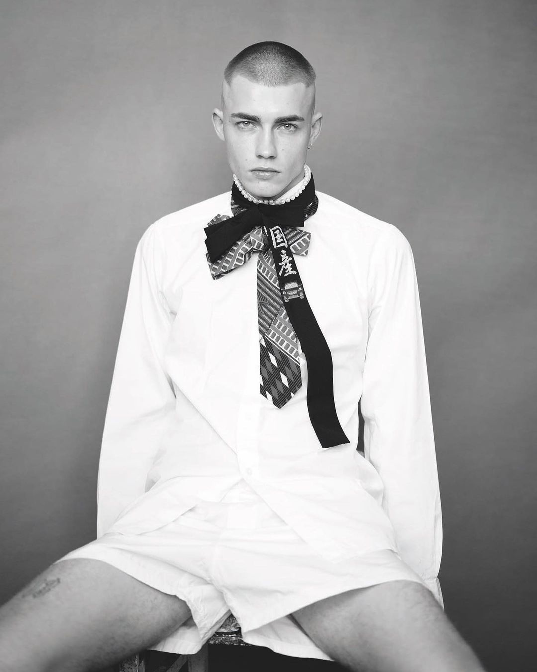 Black and white portrait of a young adult male with a buzzcut wearing a white shirt and patterned tie, seated against a gray backdrop.