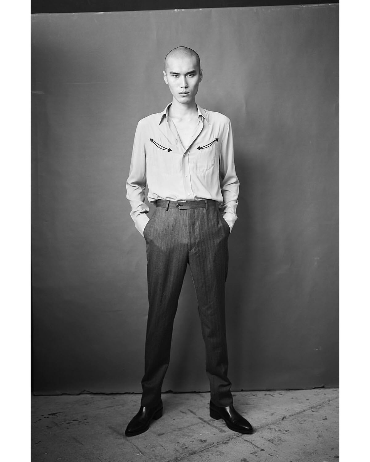 Portrait of a bald adult male in a button-down shirt and dark trousers, posing in front of a gray backdrop, standing and looking at the camera.