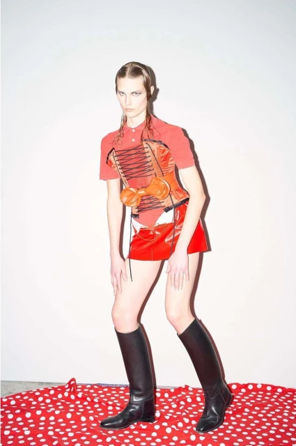 A fashion photo of a woman in red top and skirt with black boots, posing in front of a plain white wall and red and white polka dot fabric.