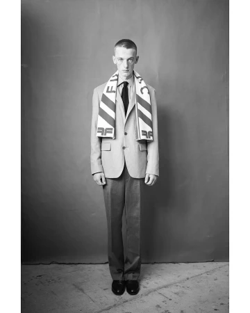 Young man in a tailored blazer and patterned scarf standing against a plain backdrop in Paris by Thomas Hauser.