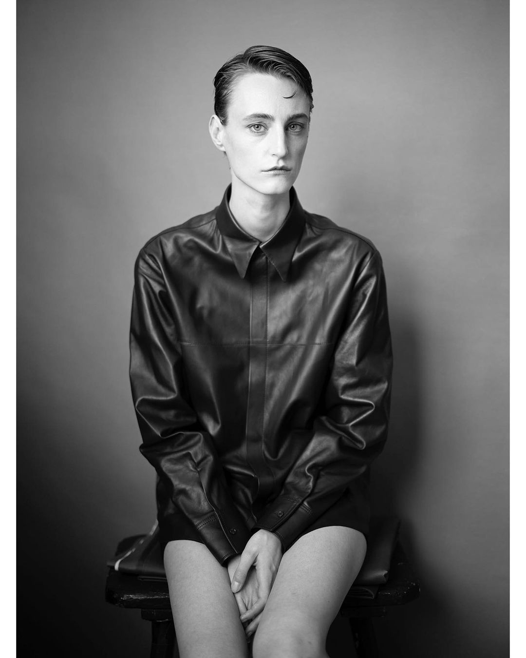Black and white portrait of a seated person with slicked-back hair and a leather-like shirt, set against a gray background in a studio.