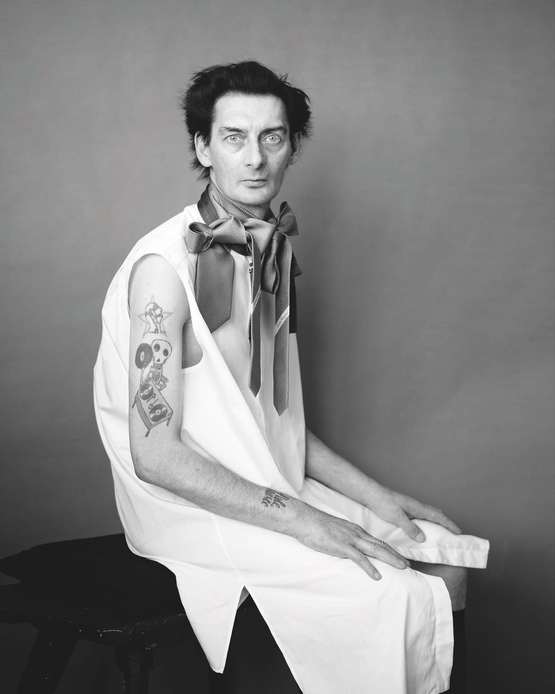 Black and white portrait of a man wearing several neckties sitting in a photo studio with a tattoo on his left arm.