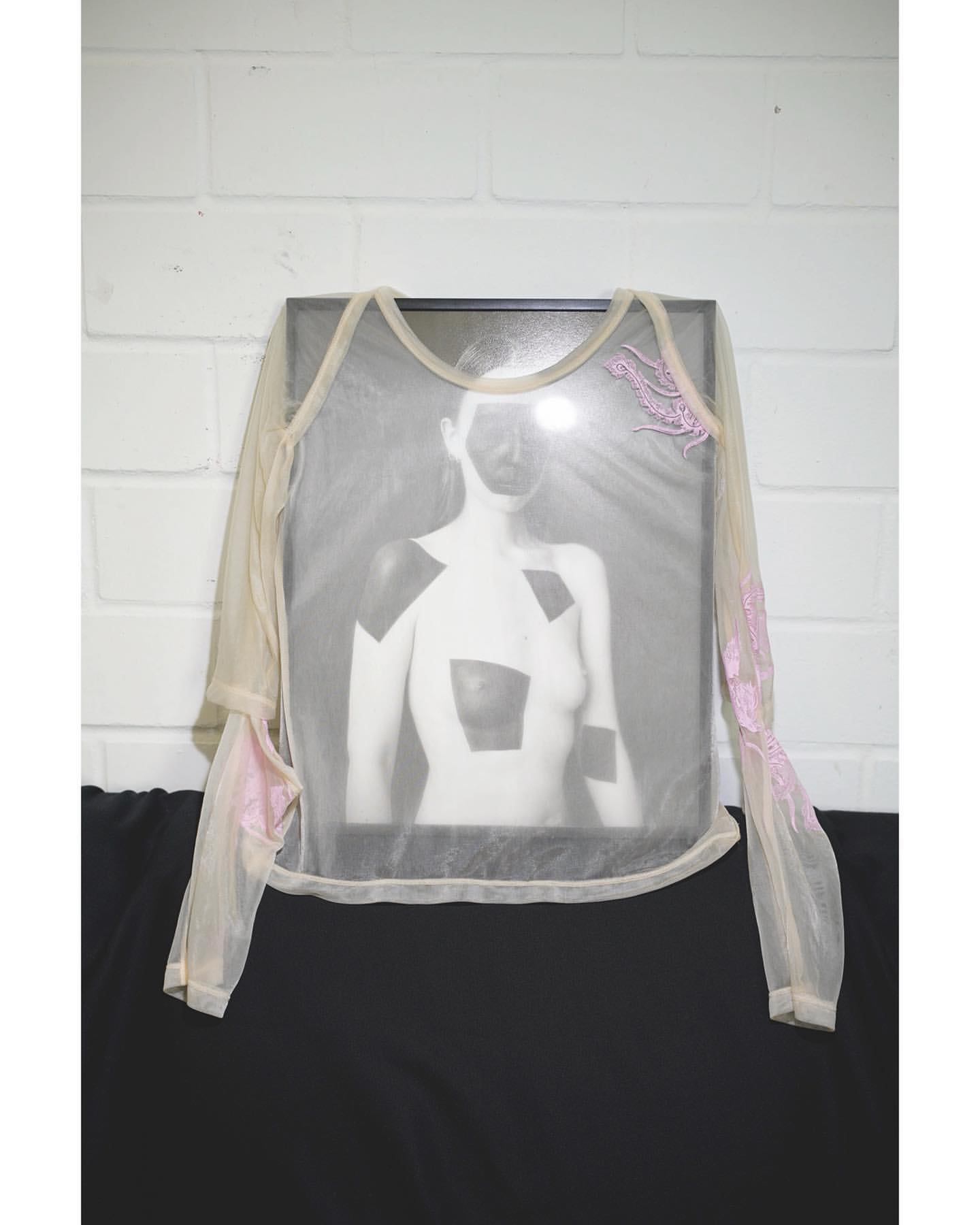 A translucent shirt with floral accents is displayed over a framed black and white photo on a black surface against a white brick wall.