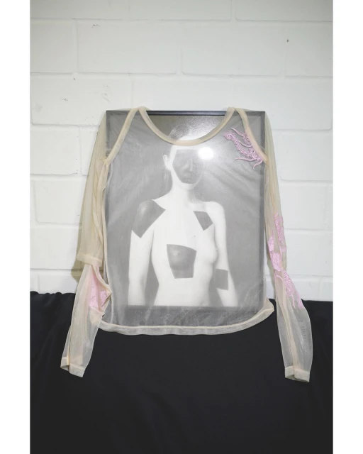 A translucent shirt with floral accents is displayed over a framed black and white photo on a black surface against a white brick wall.