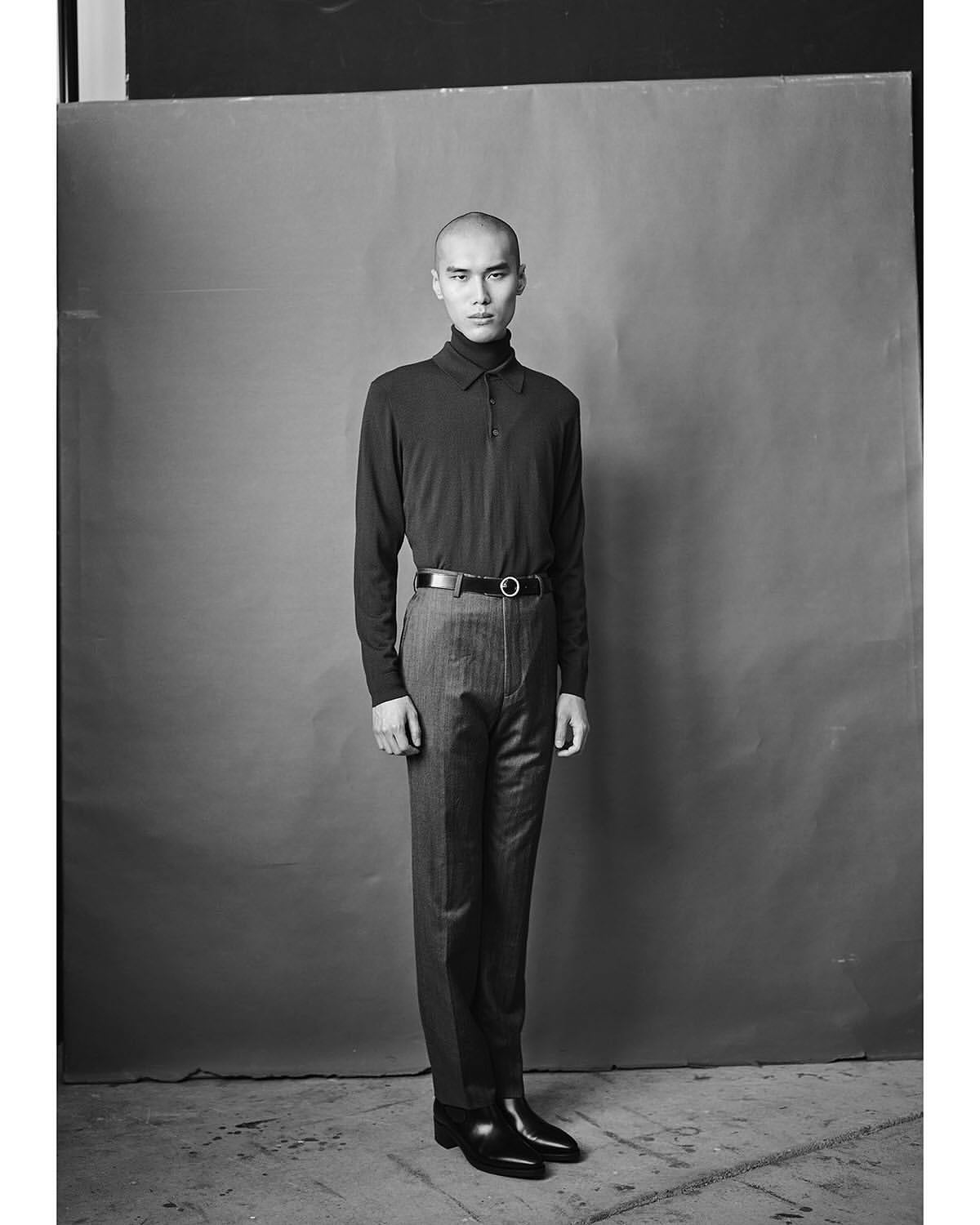 Monochrome portrait of a man standing against a seamless studio backdrop, wearing a dark collared shirt and trousers.