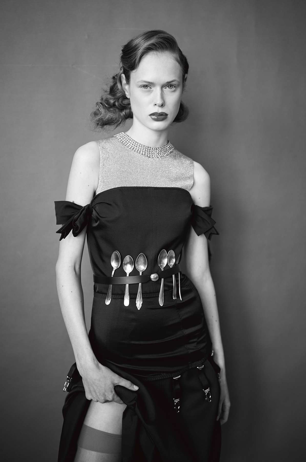 Monochrome portrait of a woman wearing a black dress and hosiery, standing in front of a plain grey background in a photo studio.
