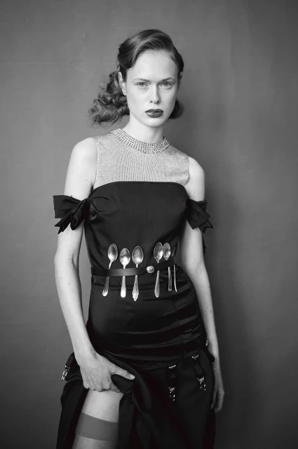 Monochrome portrait of a woman wearing a black dress and hosiery, standing in front of a plain grey background in a photo studio.