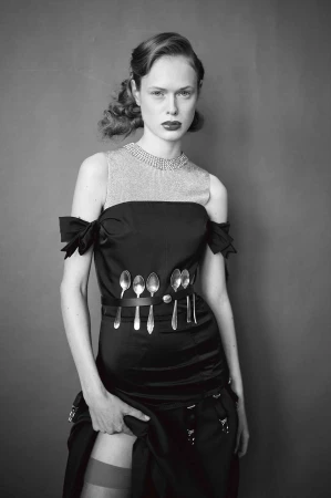 Monochrome portrait of a woman wearing a black dress and hosiery, standing in front of a plain grey background in a photo studio.