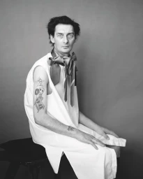Monochrome portrait of a seated man wearing a sleeveless white tunic and patterned neckties in Paris by Thomas Hauser