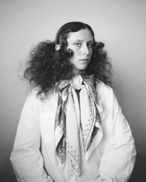 Monochrome portrait of a model with voluminous dark hair wearing a white shirt and a patterned neck scarf against a neutral background.