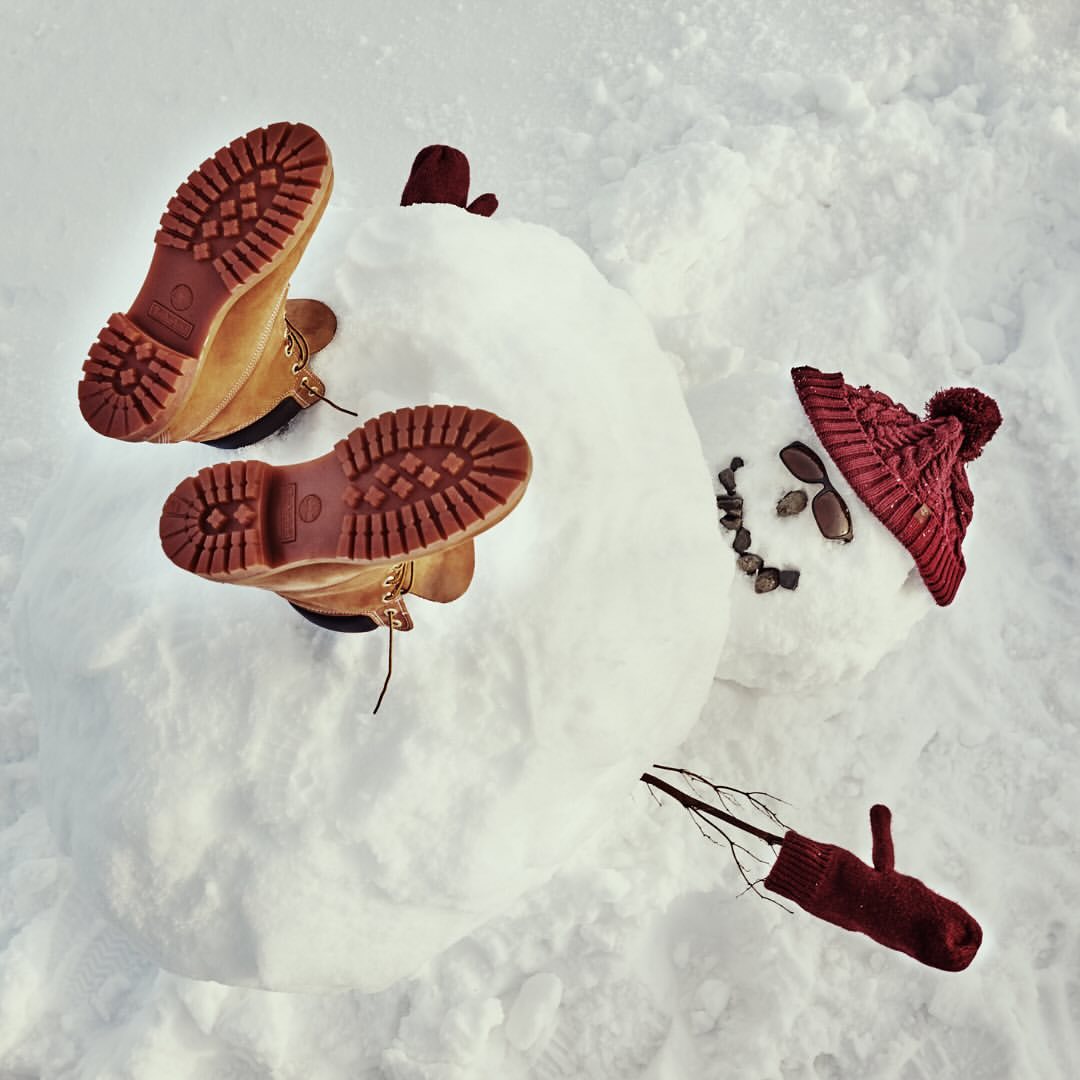 Overhead shot of a snowman with brown boots and a red hat and mittens on white snow on a sunny day in an outdoor environment.