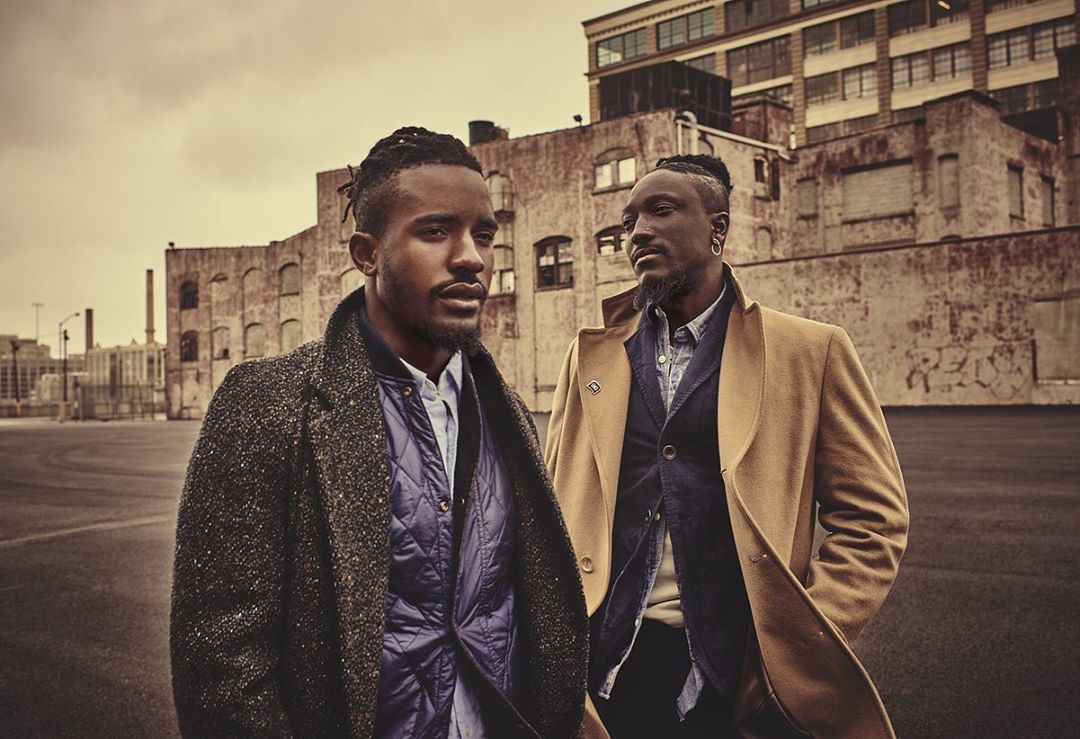 Two Black men stand outdoors in coats against a weathered brick building, capturing an urban fashion editorial look with neutral tones.