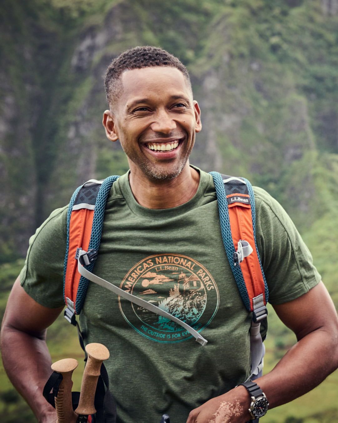 Smiling African American man wearing a backpack and holding hiking sticks, poses in a mountainous outdoor environment.