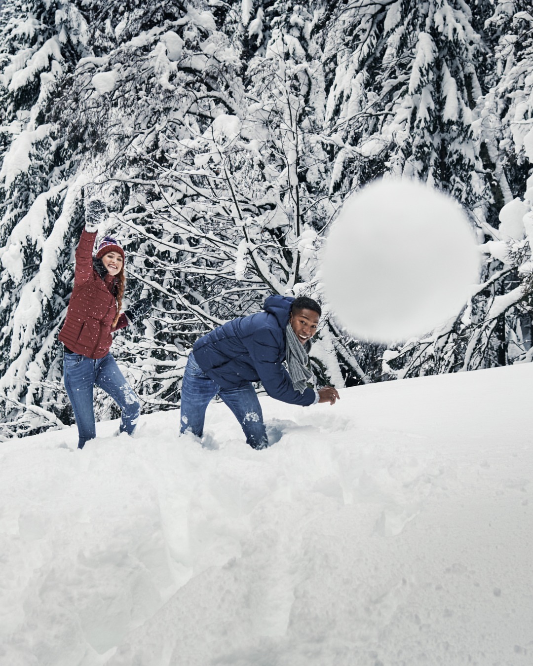 Two people, an adult male and an adult female, enjoy a playful snowball fight in a snowy, wooded environment during the winter season.
