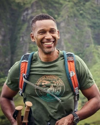 Smiling black man wearing a green graphic t-shirt and orange backpack carrying hiking poles outdoors