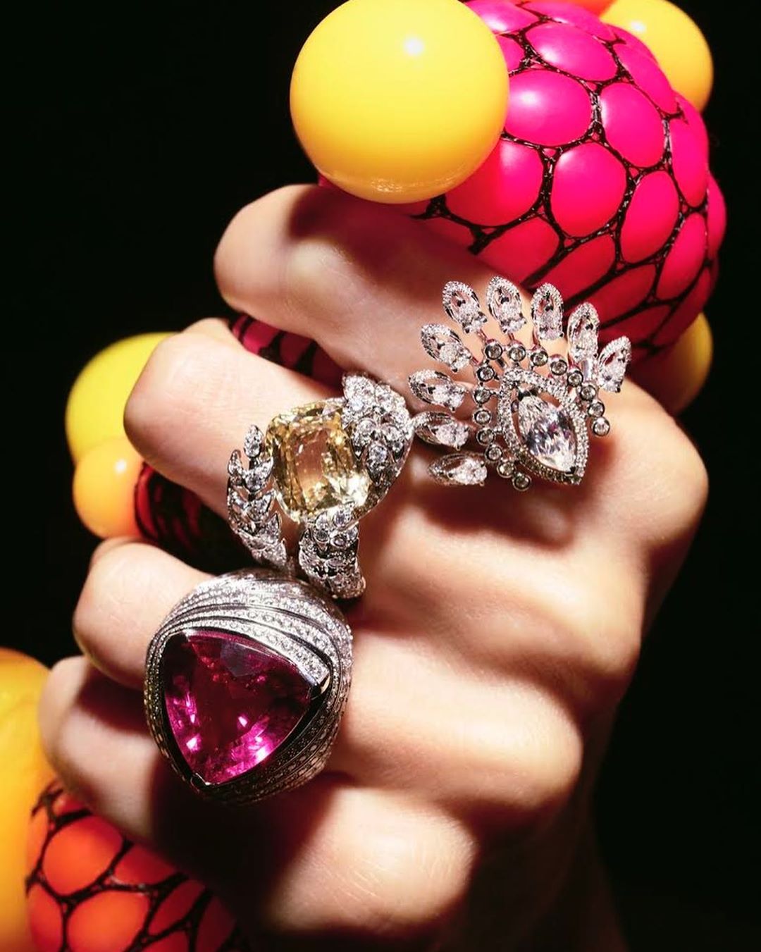 Hand displaying three cocktail rings with pink, yellow and white gems, with yellow and pink ball decorations on a black background.