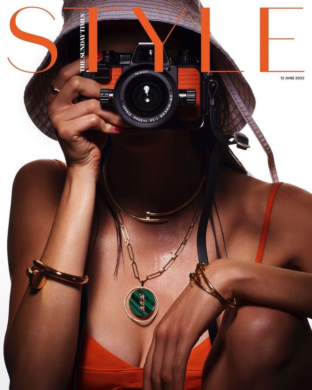 Fashion magazine cover featuring a model holding a vintage camera in front of her face, wearing jewelry and a bucket hat.