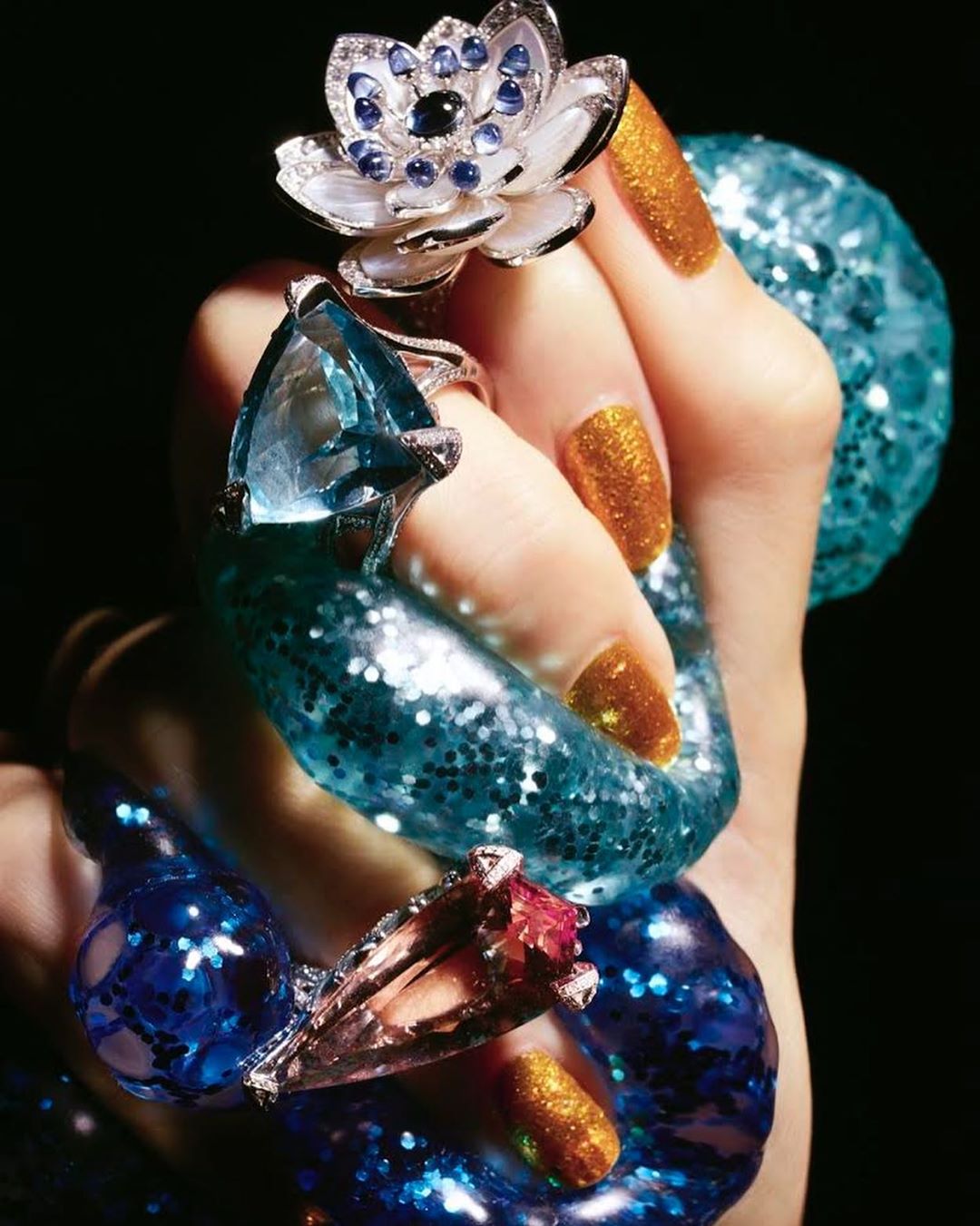 Hand holding glittery blue slime, adorned with rings, showing off golden manicure in a beauty and fashion editorial shot.