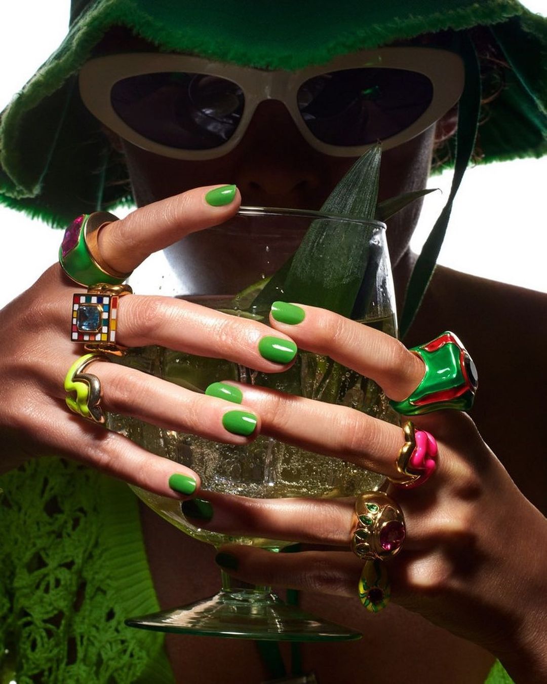 Woman in green hat and sunglasses holding a cocktail with vibrant green nails and multiple rings, set against a white background.
