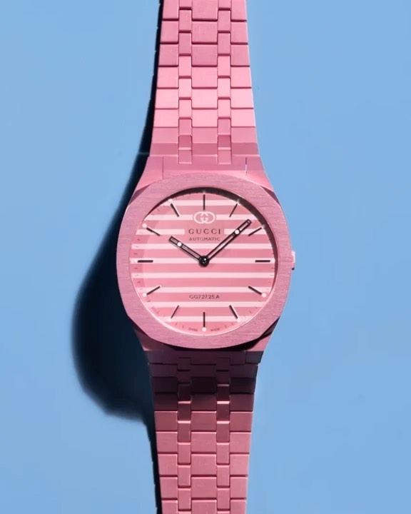Close-up studio shot of a pink Gucci watch with a striped face against a blue background, highlighting its design and color.