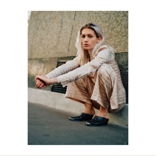 A blonde model in neutral patterned suit squats outdoors with hands clasped and wearing heeled clogs, showcasing fashion and style.