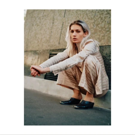A blonde model in neutral patterned suit squats outdoors with hands clasped and wearing heeled clogs, showcasing fashion and style.